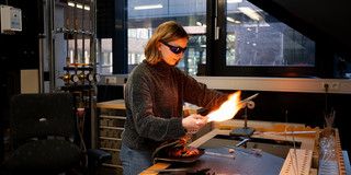 Foto: Eine Person arbeitet in einem Labor mit einer Flamme und verschiedenen Glasinstrumenten.