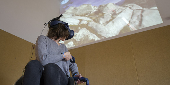 Eine Frau im Rollstuhl mit VR-Brille. Im Hintergrund eine Berglandschaft an die Wand geworfen.