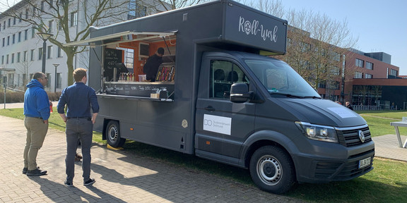 Ein dunkelgrauer Foodtruck steht auf einem Schotterweg und zwei Personen stehen davor.