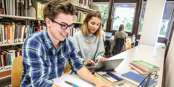 Zwei Studierende sitzen mit Lernmaterialien an einem Tisch in einer Bibliothek.