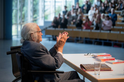 Vorne links im Profil Horst Selbiger, ein älterer Mann mit grauem Zopf und Brille im Rollstuhl, der applaudiert und lächelt. Unscharf im Hintergrund die Reihen eines Hörlesungssaals, in dem Menschen stehend applaudieren.