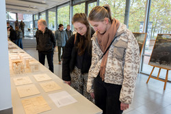 Auf dem Foto ist eine lange Tischtafel mit Modellen aus Holz zu sehen. Besucher*innen schauen sich diese an. Im Hintergrund ist eine helle Fensterfront zu sehen und Bilder auf Bildstaffeleien.