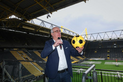 Rektor Manfred Bayer, der ein Mikrofon und einen gelb/schwarzen Fußball in der Hand hält. Im Hintergrund das Fußballfeld und die leeren Tribünen des BVB-Stadions.