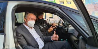 Zwei Männer sitzen in einem Auto. Bei tragen eine Maske. Der Mann auf dem Beifahrersitz zeigt den Daumen nach oben.