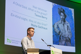 Ein Sprecher an einem Vortragspult, eine PowerPoint-Präsentation im Hintergrund
