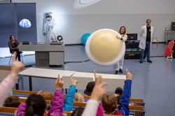 Eine Frau steht in einem vollbesetzten Hörsaal und hält einen großen Wasserball, der den Planeten Saturn symbolisiert, in der Hand. Im Publikum sitzen Kinder und zeigen auf.