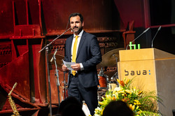 Ein Mann mit kurzen, dunklen Haaren im Anzug steht auf einer Bühne und spricht.