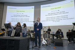 Ein Sprecher und eine Band stehen in einem Hörsaal vor einer Leinwand mit dem Text des Liedes Feliz Navidad.