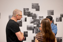Drei Personen stehen vor einer Wand, auf der viele kleine Bilder in schwarz-weiß angebracht sind