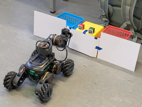 Spielerisch zur assistiven Robotik Ein vierbeiniger Roboter mit Kamera steht vor einer Versuchsaufstellung mit drei farbigen Körben und markierten Würfeln hinter einer Papierbarriere.