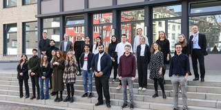 Eine Gruppe von Frauen und Männern stehen auf Treppenstufen vor einem Gebäude.