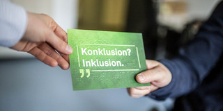 Foto: Zwei Personen reichen sich eine grüne Karte mit der Aufschrift „Konklusion? Inklusion.“ und Blindenschrift darauf.