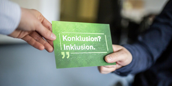 Foto: Zwei Personen reichen sich eine grüne Karte mit der Aufschrift „Konklusion? Inklusion.“ und Blindenschrift darauf.