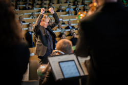 Auf dem Foto ist der Dirigent zu sehen, wie er das Orchester leitet.