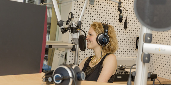 A woman with headphones sits in the studio and speaks into a microphone