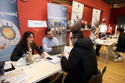 Infostand der Firma ICA Traffic GmbH auf der Internationalen Karrieremesse Ein Mann sitzt an einem Tisch. Ihm gegenüber sitzen eine Frau und ein Mann, der einen Laptop vor sich stehen hat. Im Hintergrund sind zwei Aufsteller der Firma ICA Traffic GmbH zu sehen.