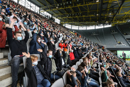 Erstsemesterstudierende, die auf einer Tribüne in einem Fußballstadion mit Mund-Nasen-Bedeckung sitzen, reißen ihre beiden Arme hoch und machen eine La-Ola-Welle.