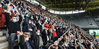 Erstsemesterstudierende, die auf einer Tribüne in einem Fußballstadion mit Mund-Nasen-Bedeckung sitzen, reißen ihre beiden Arme hoch und machen eine La-Ola-Welle.