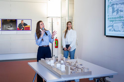 Das Bild zeigt ein Architekturmodell aus Holz, welches auf einem weißen Tisch steht. Dahinter stehen zwei weibliche Personen. Eine von ihnen hält ein Mikrophon in der Hand und schaut in Richtung einer Präsentation, die auf dem Bild nur abgeschnitten zu erkennen ist. Im Hintergrund der beiden Frauen ist eine Tür sowie eine weiße Wand mit Fotos zu sehen.
