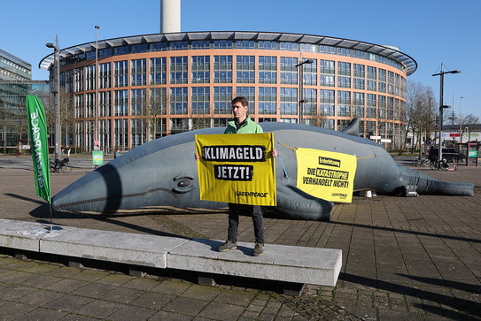Foto: Eine Person hält Protestschilder vor einem großen Gebäude mit einem aufblasbaren Wal im Hintergrund.