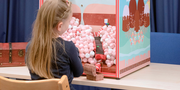 Back view of a girl sitting in front of a box. In the box you can see a staircase made of chocolate, which leads to a treasure box. 