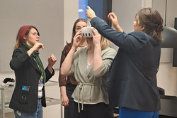 Das Bild zeigt eine weiblich gelesene Person mit einer VR-Brille. Weitere weiblich gelesen Personen stehen um sie herum. Im Hintergrund ist ein Teil eines Fensters zu sehen.