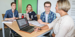 Zwei Studentinnen und zwei Studenten im Gespräch. Auf dem Tisch sind Laptops und Bücher zu sehen.