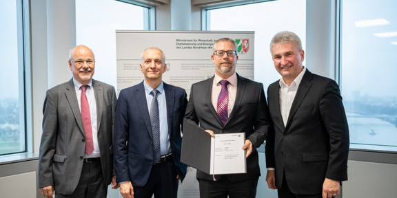 Four men in suits are standing next to each other. One of them has a document in his hand.