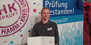 Foto: Die Chemielaborantin Bianca Beck hält ihre Auszeichnung für einen Sehr-Guten-Abschluss vor einem Poster der IHK in die Höhe.
