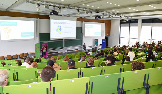 Auftaktveranstaltung des Westenergie Manager Cups 2023 Blick nach vorne zur Tafel von den oberen Reihen eines Hörsaals, in dem Studierende einer Präsentation lauschen.