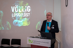 Das Bild zeigt Rektor Bayer hinter einem Rednerpult mit Mikrofon in der Hand. Hinter ihm an der Wand ist eine Präsentation mit dem Titel "Völlig losgelöst" zu sehen.