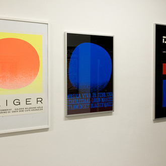 Drei Künstlerplakate hängen an einer weißen Wand nebeneinander. Die rechten haben dunkelblauen und schwarzen Hintergrund. 