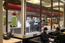 Hinter Fensterglas ist ein Raum mit vielen Gerätschaften wie etwa Druckern zu erkennen. Vor dem Fensterglas sitzen zwei Studierende an ihren Laptops.