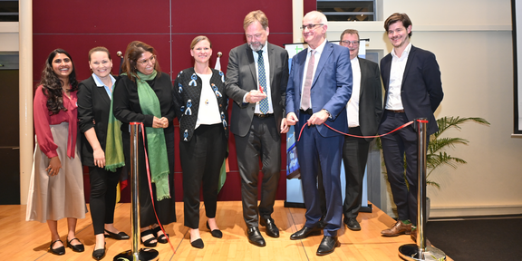 Foto: Eine Gruppe von Menschen steht auf einer Bühne und schaut dabei zu wie ein Mann in einem Anzug mit einer Schere ein rotes Band durchtrennt.