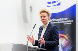 A woman with blond hair stands at the lectern in front of a UA Ruhr poster.