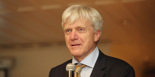 Foto. Porträtfoto von Andreas von Bechtolsheim vor braunem Hintergrund. Er ist ein Mann im Anzug mit weißen Haaren und Krawatte.