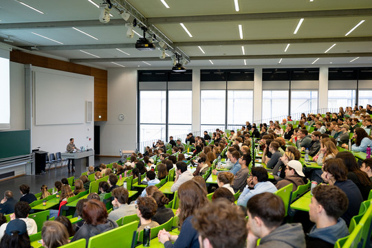 Auf dem Foto sieht man einen vollen Hörsaal mit Studierenden und einem Gastredner vorne sprechen.