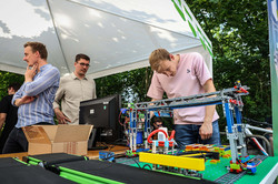 Ein Mann betrachtet eine Installation aus Lego-Steinen und Kabeln. Links stehen zwei weitere Männer.