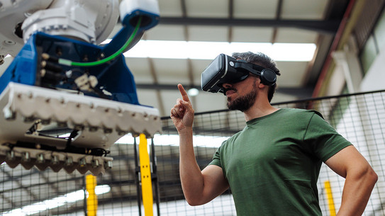 Foto: Eine Person trägt eine VR-Brille und interagiert mit einem großen Industrieroboter in einer Werkhalle.