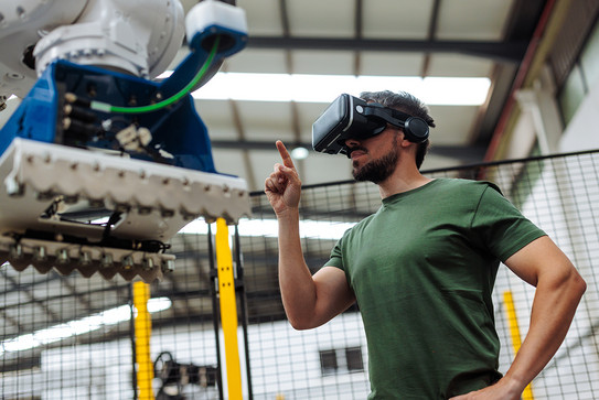 Foto: Eine Person trägt eine VR-Brille und interagiert mit einem großen Industrieroboter in einer Werkhalle.