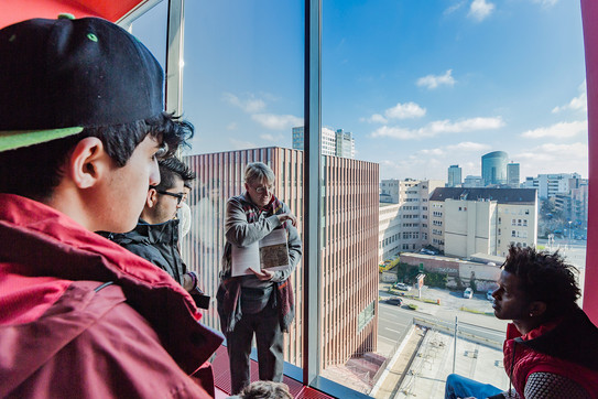 Prof. Barbara Welzel steht mit mehreren Studenten vor einer großen Fensterscheibe im oberen Stockwerk eines Gebäudes. Das Fenster bietet eine Aussicht auf viele Gebäude.
