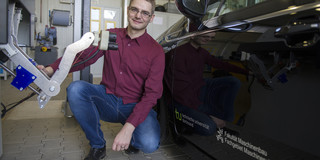 Man in dark red shirt kneels between black car and robot arm