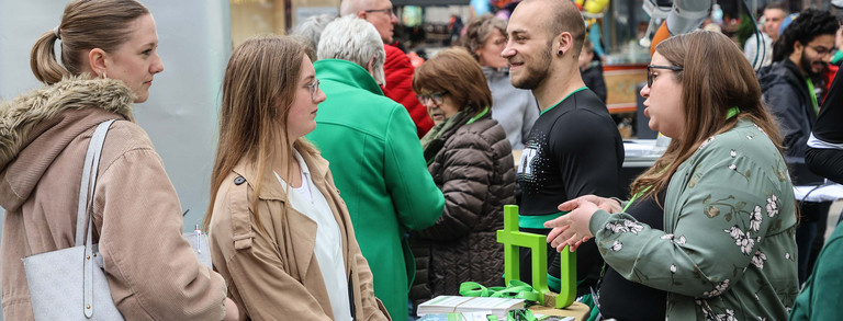 Foto: Bei einem Stadtfest in der Dortmunder Innenstadt steht eine Frau mit Brille an einem Stand der TU Dortmund und berät zwei junge Frauen. Im Hintergrund sieht man Luftballons und einen Roboter.