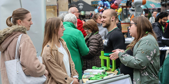 Foto: Bei einem Stadtfest in der Dortmunder Innenstadt steht eine Frau mit Brille an einem Stand der TU Dortmund und berät zwei junge Frauen. Im Hintergrund sieht man Luftballons und einen Roboter.