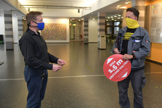 Christian Bartsch und Frank Kornhof stehen sich mit Mundschutz und Abstandsschild gegenüber.