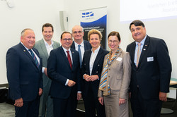 A group of men and women in suits smile at the camera in front of a UA Ruhr poster.