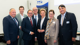 Eine Gruppe von Männern und Frauen in Anzügen lächeln vor einem Poster der UA Ruhr in die Kamera..