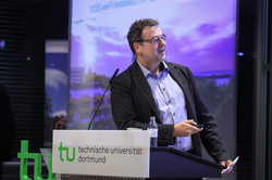 Foto: Eine Person steht am Rednerpult der Technischen Universität Dortmund und zeigt auf eine Präsentation im Hintergrund.