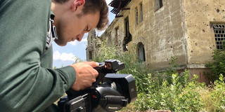 A man holds a camera pointed at a dilapidated building