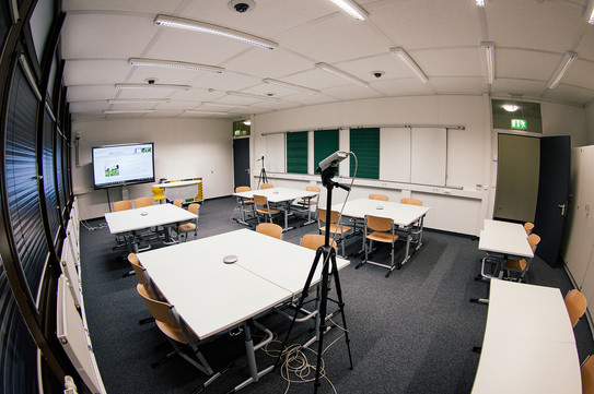 Innenansicht eines Unterrichtsraumes, in dem mehrere Tischgruppen stehen. An einem Ende des Raumes hängt ein Monitor. 